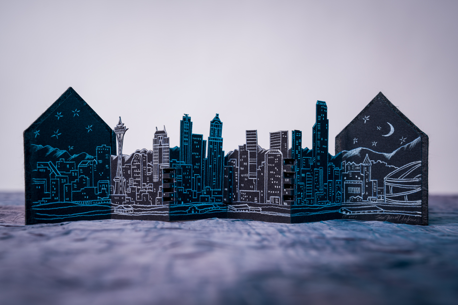An image of Seattle Skyline by Sam Garriott Antonacci against a grey-white background opened in an accordian style. The pages have an alternating blue/white pattern and are cut to look like a city skyline.